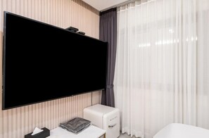 A hotel room with a large flat-screen TV mounted on the wall, a small white bedside table with a tissue box and a magazine, a white bedside unit, and sheer white curtains. - Hotel Yaja Gwangju Mudeungsan branch (Gwangju)
