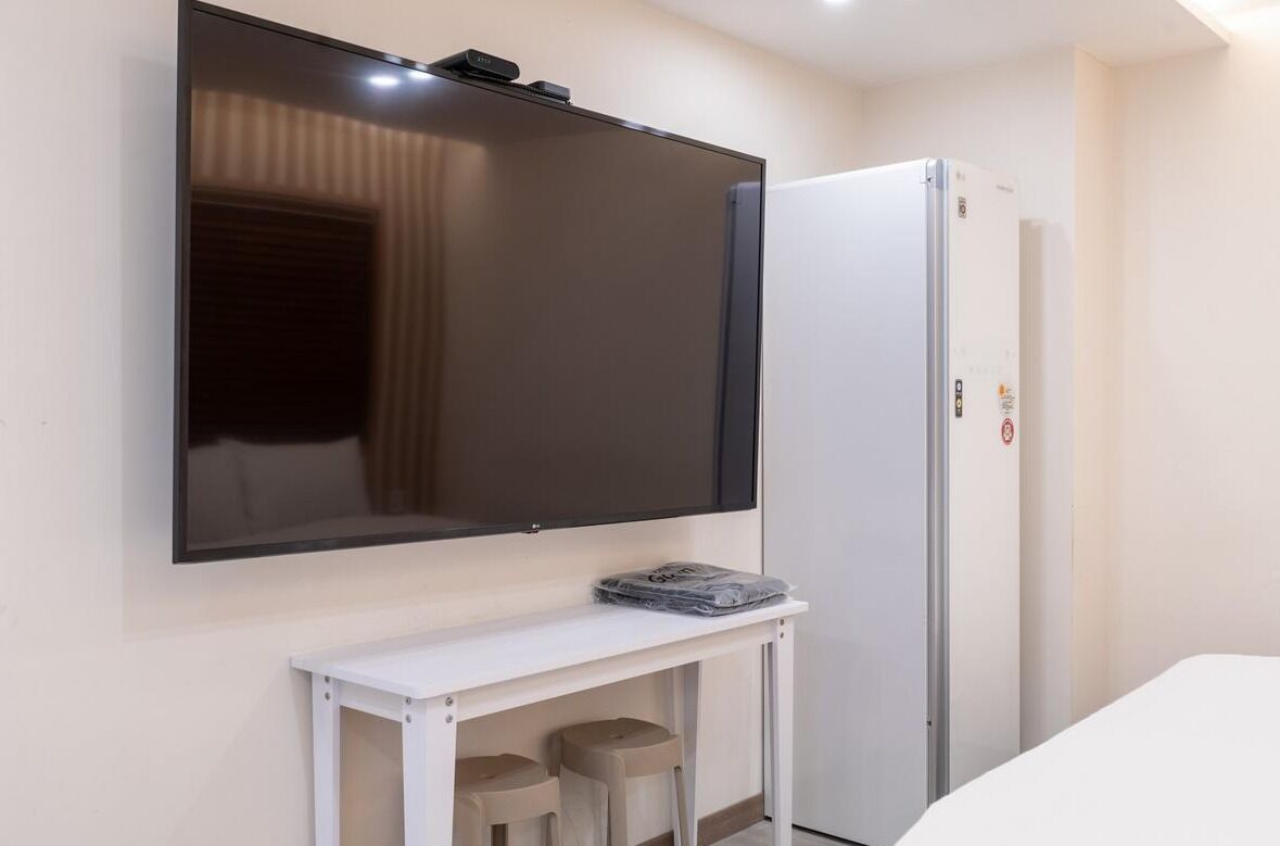 A hotel room with a flat-screen TV mounted on the wall, a white bedside table with a remote control, and a white wardrobe.