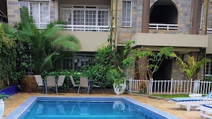 Piscina com espreguiçadeiras, rodeada por vegetação e um edifício com varandas.