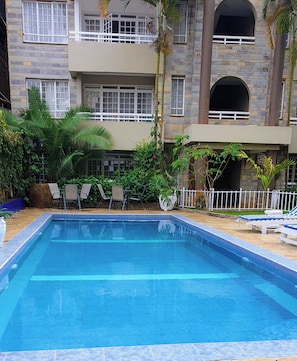 Una piscina con sillas de descanso, rodeada de follaje y un edificio de varios pisos con balcones.