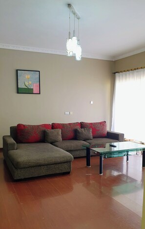 A living room with a sectional sofa, a glass coffee table, a pendant light, a framed picture on the wall, and a window with curtains. - Luxurious 2 Bedroom in Kilimani With Scenic Views and Modern Amenities (Nairobi)