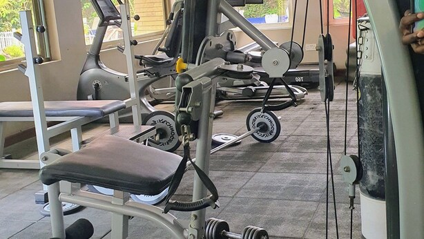 Uma academia com diversos equipamentos de ginástica, janelas amplas e vista para prédios.