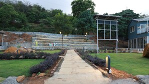 A stone pathway leading to a seating area with a stone wall backdrop, surrounded by greenery and a building with a balcony. - Harmony Hills (Paud)