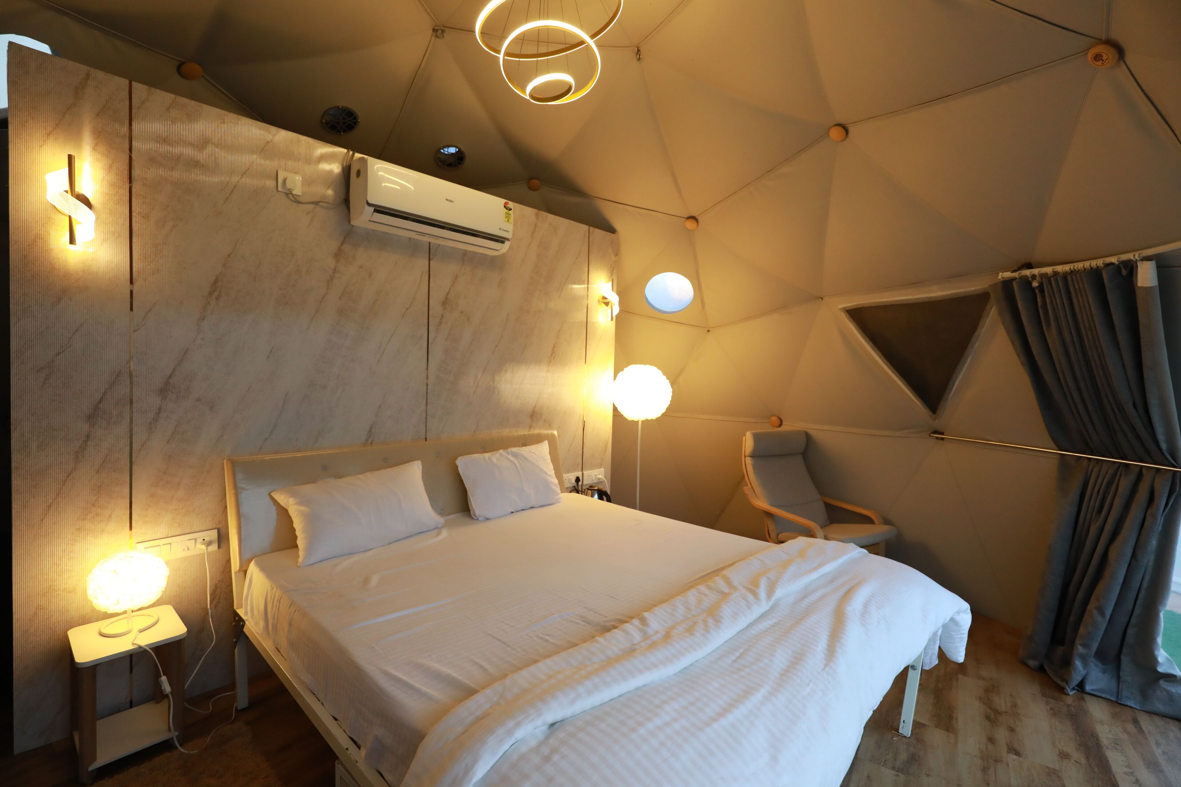 Ein modernes Hotelzimmer mit einem großen Bett, einem Schreibtisch und einem Sessel. An der Wand befindet sich eine Klimaanlage und ein Fenster mit Vorhängen.