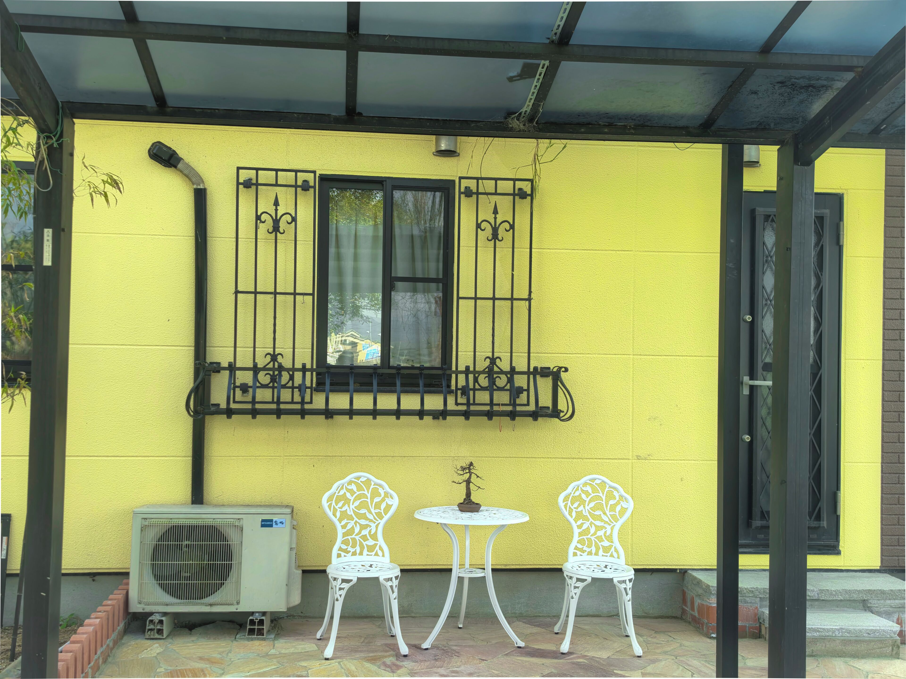 A yellow building with a window, two white chairs, and an air conditioning unit.