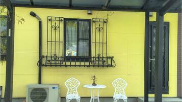 A yellow building with a window, two white chairs, and an air conditioning unit.