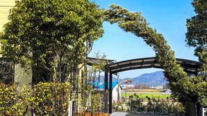 A gated entrance with a covered walkway, a building with a blue roof, and a view of distant mountains. - Onsengunyado (Toon)