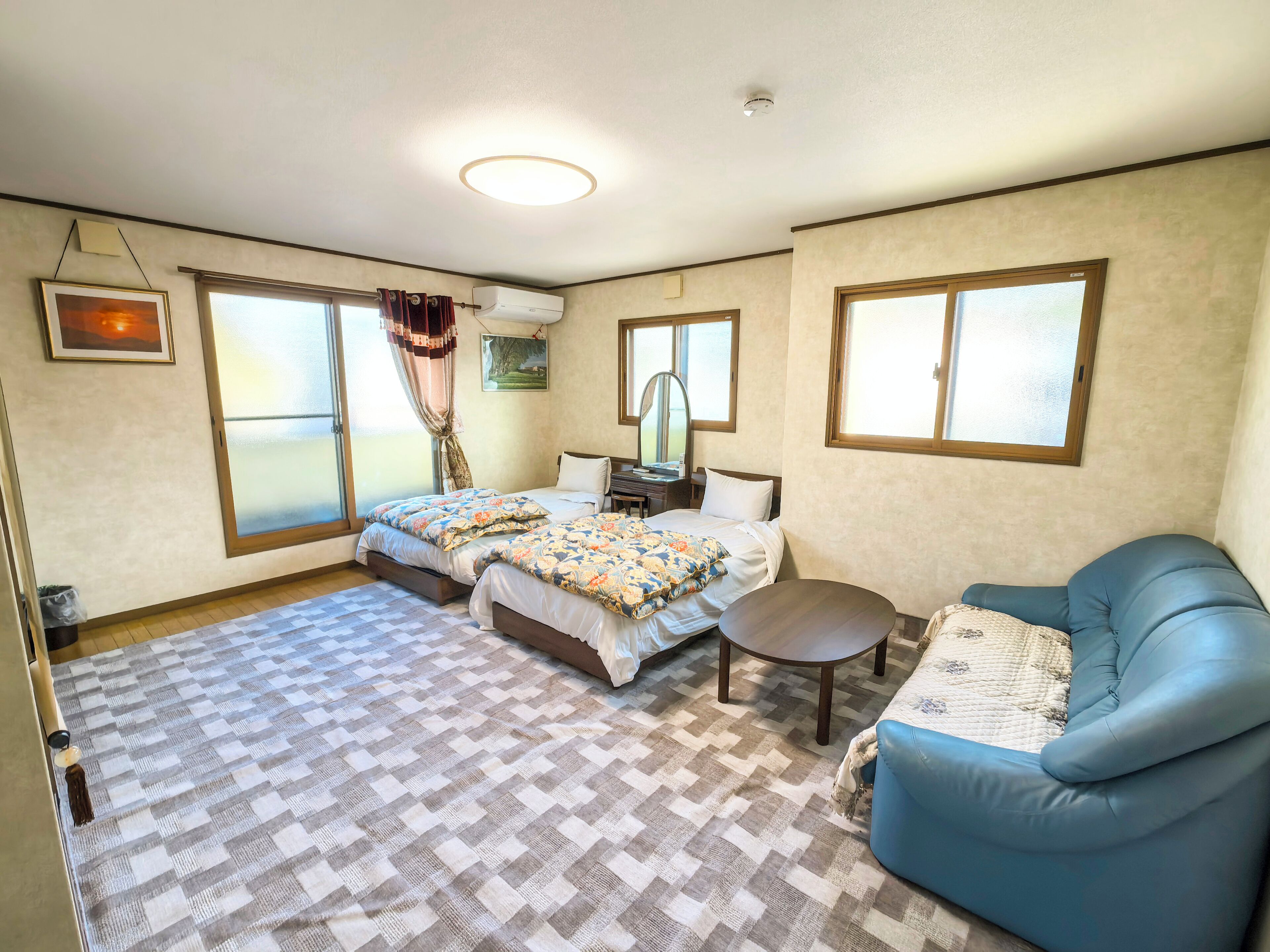 A hotel room with two beds, a blue sofa, a small table, and large windows.