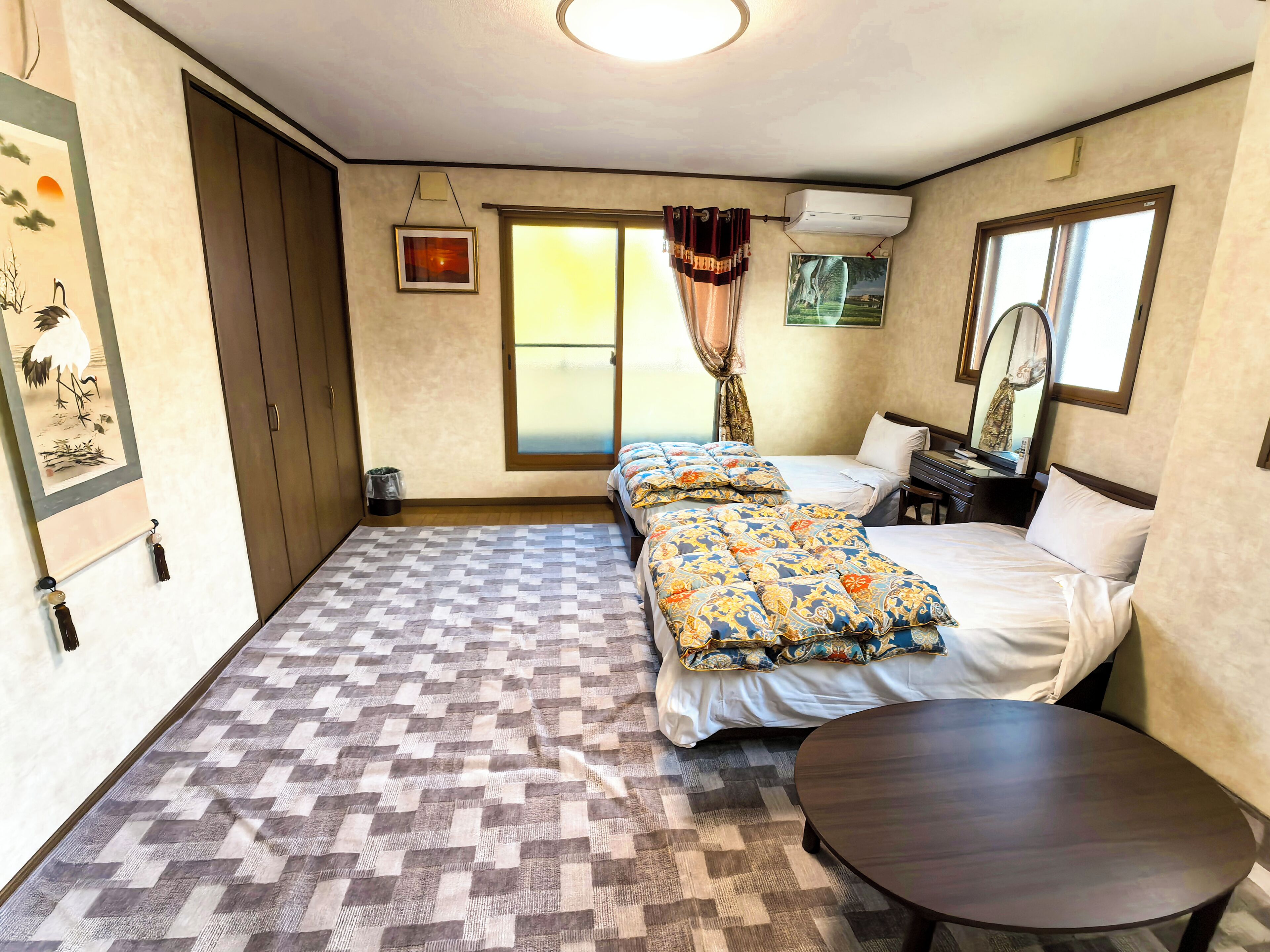 Un dormitorio con suelo a cuadros, una cama con ropa de cama estampada, una pequeña mesa redonda y una ventana con cortinas.