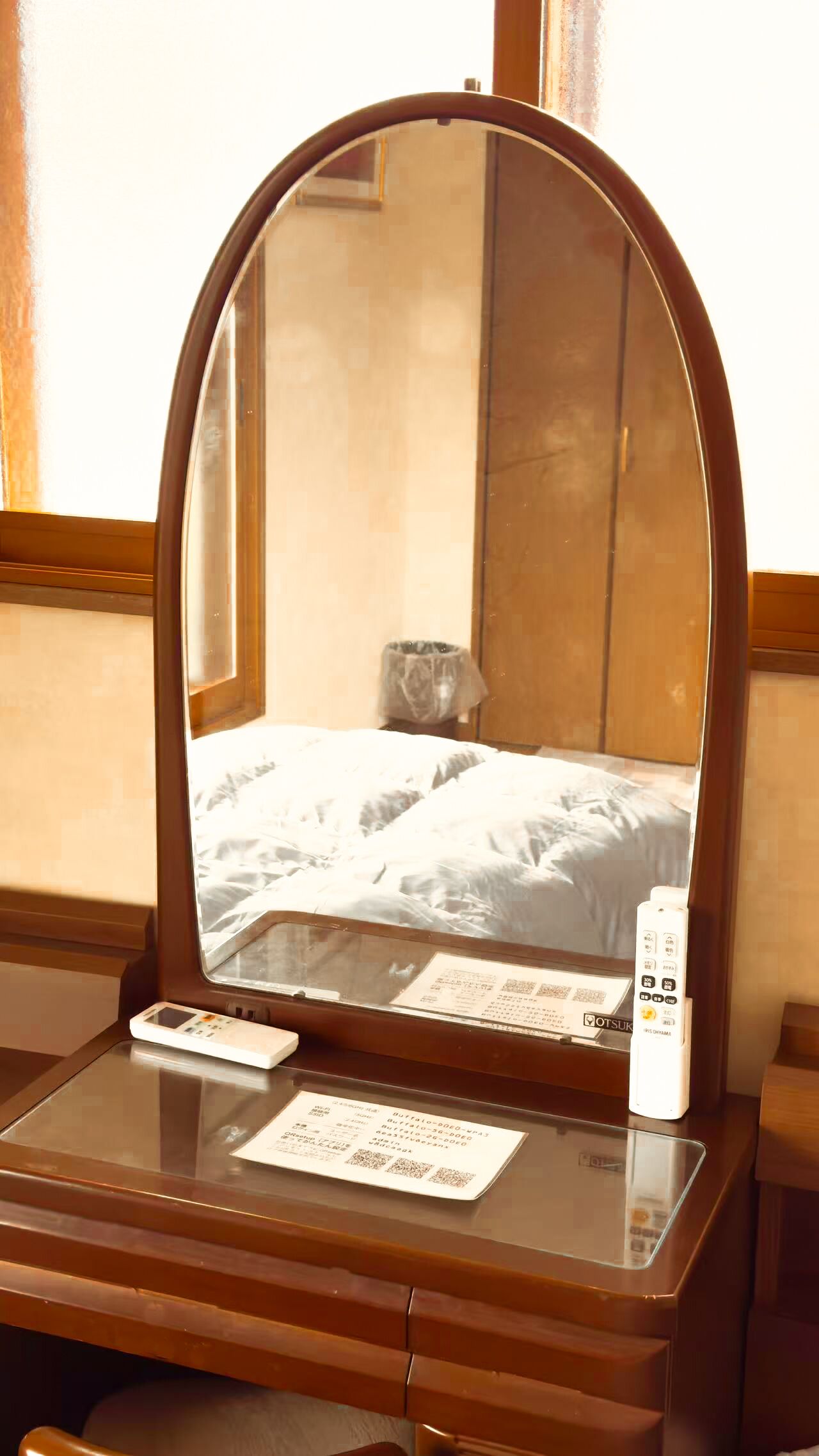 A hotel room with a wooden vanity, a mirror, a bed with white bedding, a bedside table with a remote control, and a window with wooden frames.