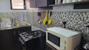 Een keuken met een gasfornuis, magnetron, betegelde spatwand, hangende keukengerei en een raam boven de gootsteen.