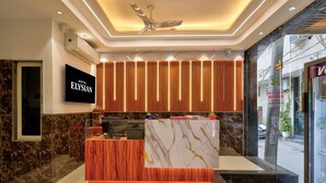 A hotel reception area with a marble counter, wooden paneling, a ceiling fan, and a TV screen displaying the hotel name. - Hotel Elysian at Delhi Airport  (New Delhi)