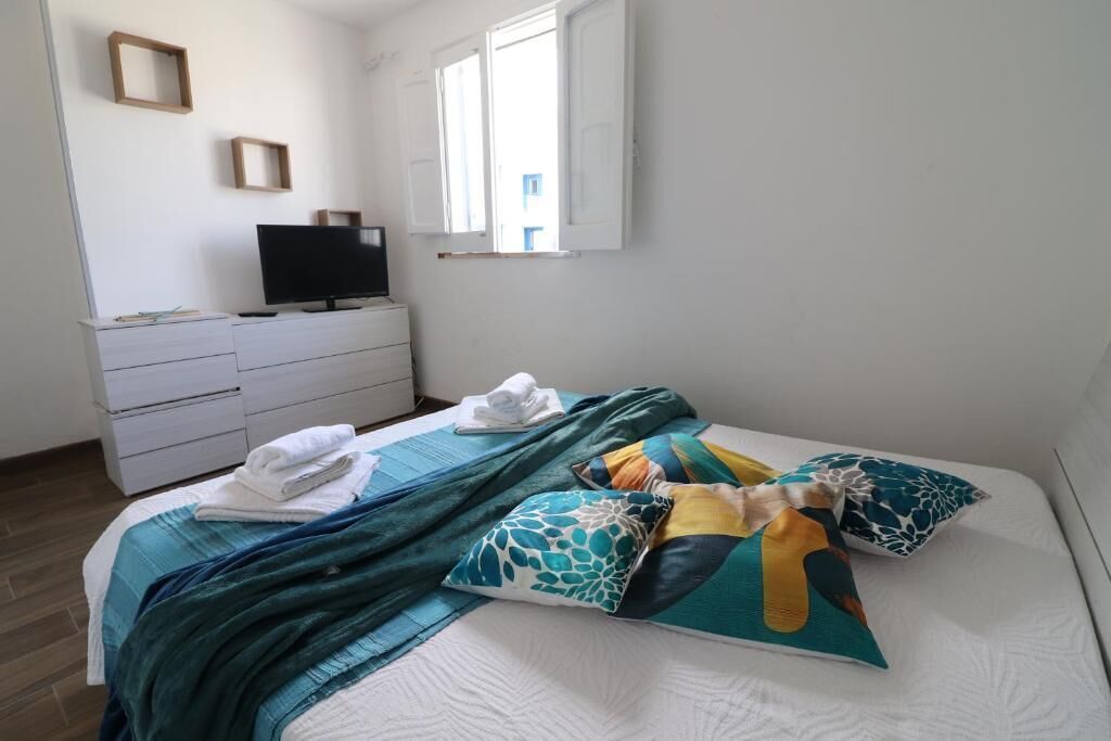 Un dormitorio con cama, televisor en una cómoda, ventana y pared blanca.