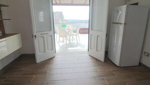 Una habitación con piso de madera, un refrigerador blanco, una puerta blanca que da a un balcón con mesa y sillas, y vista al mar.