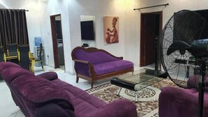 A living room with a purple sofa, a glass coffee table, a large fan, and a painting on the wall. - Charming Executive Suite with Garden Views at AJALA Lodge, Badagry (Badagry)