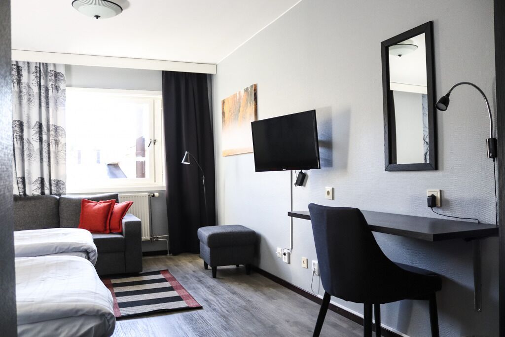 A modern hotel room with a bed, a desk, a chair, a TV, and a window with curtains.