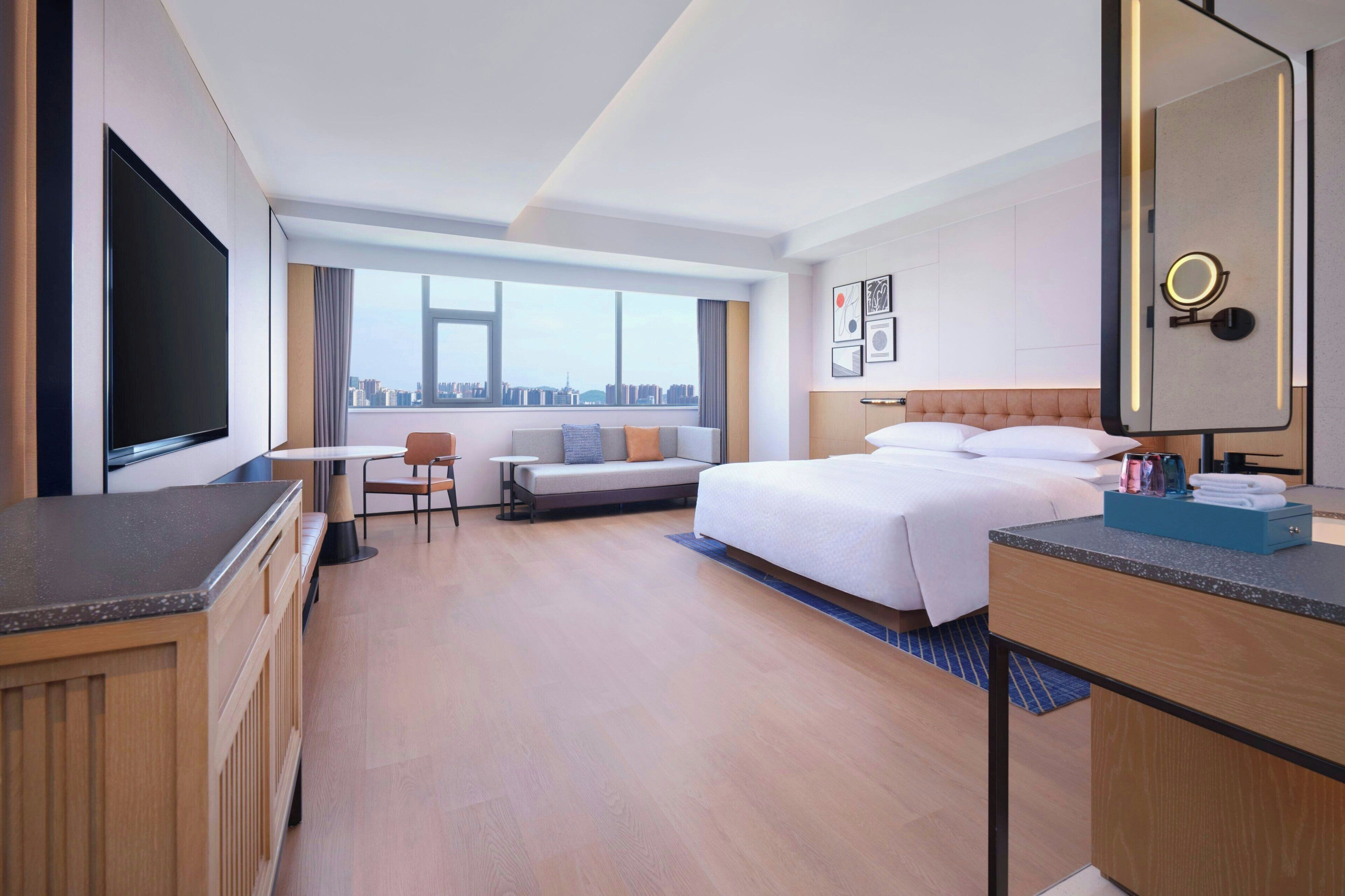 Ein Hotelzimmer mit Bett, Schreibtisch, Stuhl, Fernseher und einem großen Fenster mit Blick auf die Stadt.