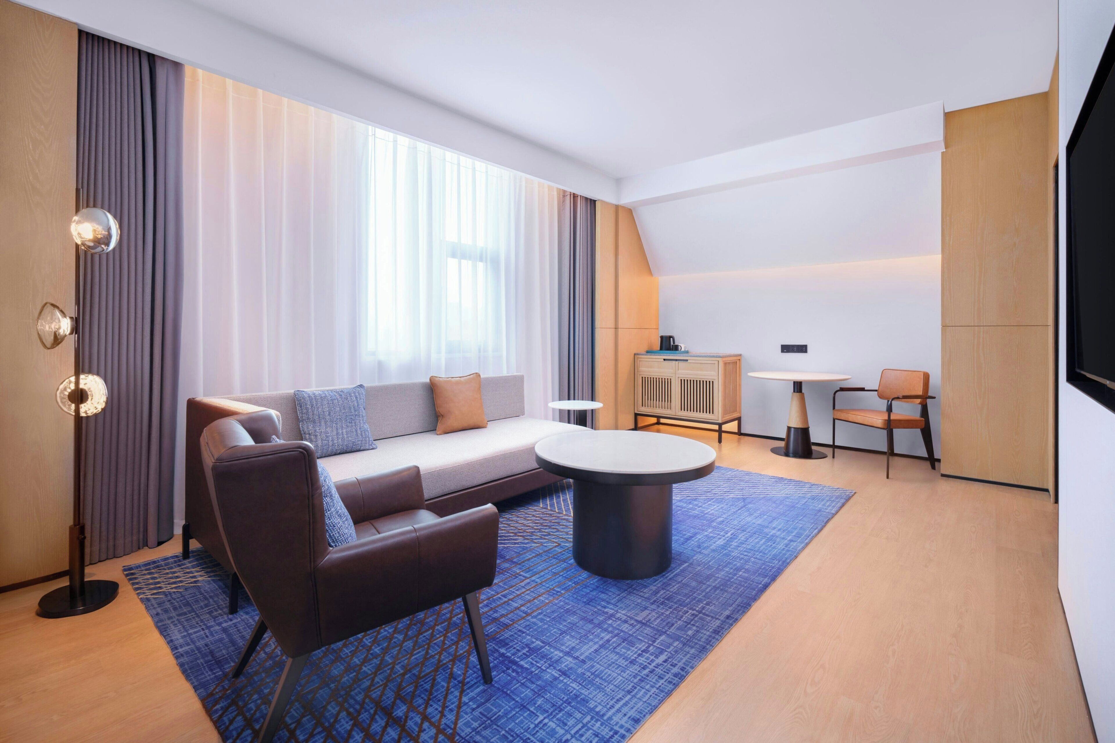 A modern hotel room with a sectional sofa, a round coffee table, a small dining table, and a flat-screen TV mounted on the wall.