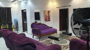 Una sala de estar con un sofá morado, una mesa de centro de vidrio, un ventilador grande y un cuadro en la pared.