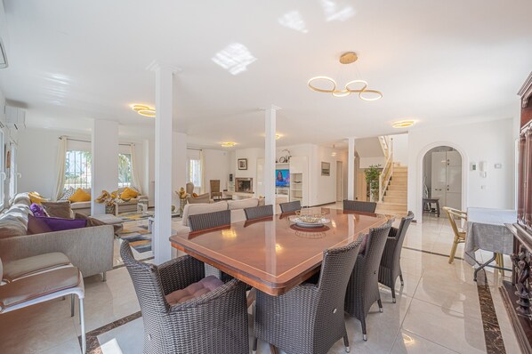 A spacious living area with a large wooden dining table surrounded by wicker chairs, a sofa set, and a staircase leading to an upper floor.