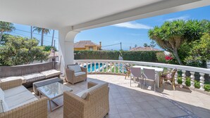 Un patio techado al aire libre con mobiliario de mimbre, una mesa de vidrio y vistas a una zona residencial con piscina.