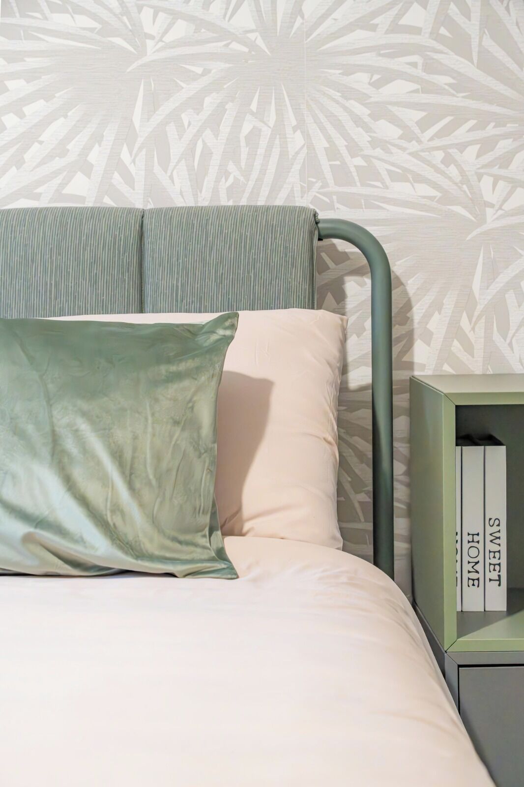 A bed with a green metal frame, a green pillow, and a light-colored mattress. There is a green nightstand with a book titled 'SWEET HOME' beside the bed.