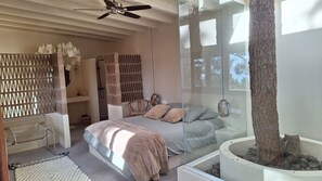 A bedroom with a bed, a chair, a bedside table, a glass partition, and a ceiling fan.