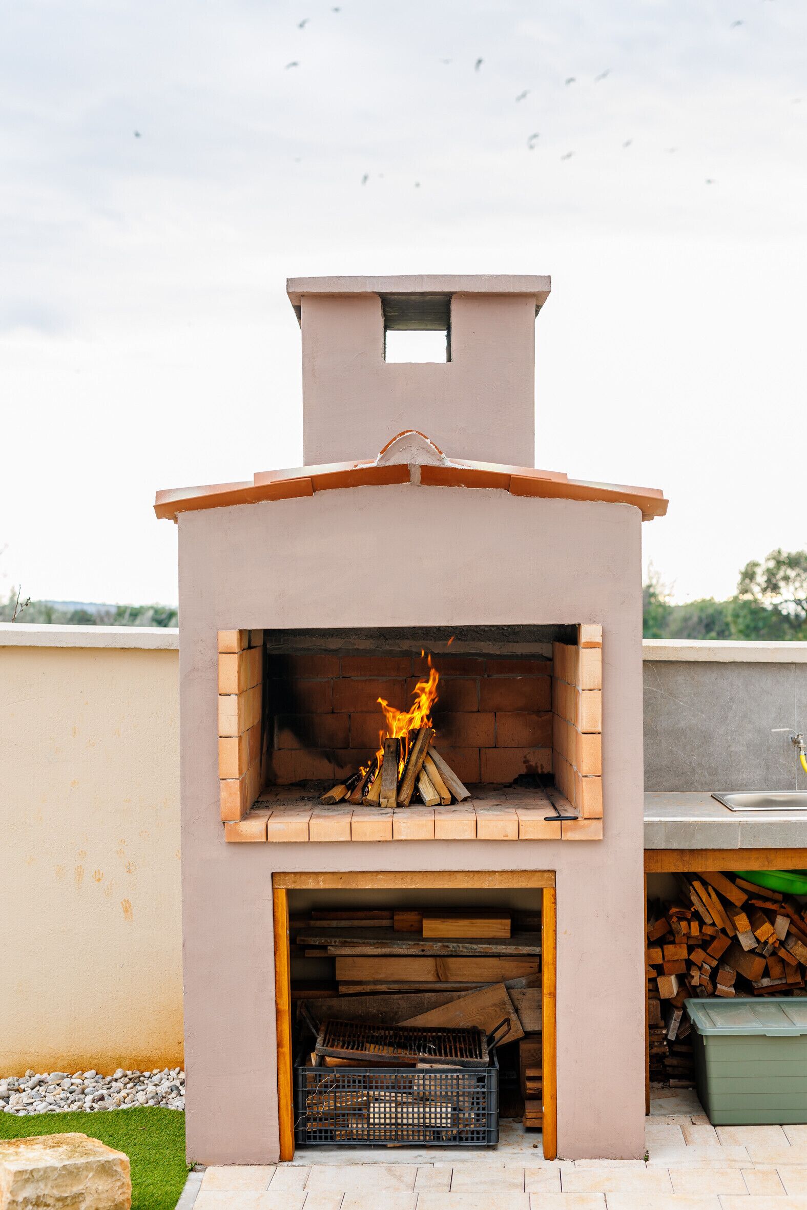 Un horno de ladrillo al aire libre con fuego encendido en su interior, una pila de leña y un contenedor verde cercano.
