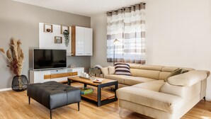 A modern living room with a sofa, a black ottoman, a wooden coffee table, a TV, and a decorative plant. - Holiday Home Fulia with pool (Galižana)