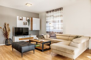 Ein modernes Wohnzimmer mit einer Couch, einem schwarzen Hocker, einem Holztisch, einem Fernseher und einer Zimmerpflanze.