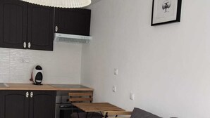 Un salon moderne avec un canapé gris, une petite table ronde, un coin cuisine doté de meubles sombres, un ventilateur de plafond blanc et un tableau encadré accroché au mur.