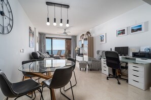 A modern living room with a glass table, black chairs, a sofa, a desk with a computer, and a ceiling fan.