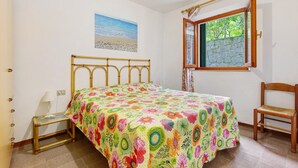 Un dormitorio con una cama, una silla, un ventanal con vistas a zonas verdes y un cuadro colgado en la pared.