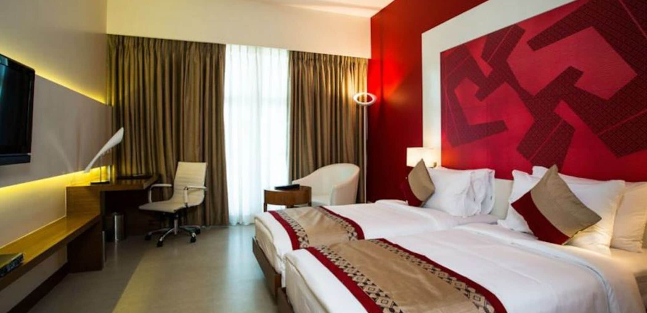 Ein Hotelzimmer mit zwei Betten, einem Schreibtisch, einem Stuhl, einem Fernseher und einem großen Fenster mit Vorhängen.