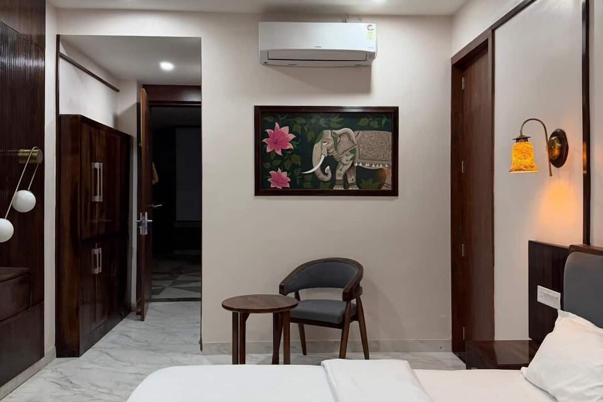 A hotel room with a bed, a chair, a small table, an air conditioner, a ceiling fan, and a wall-mounted lamp.