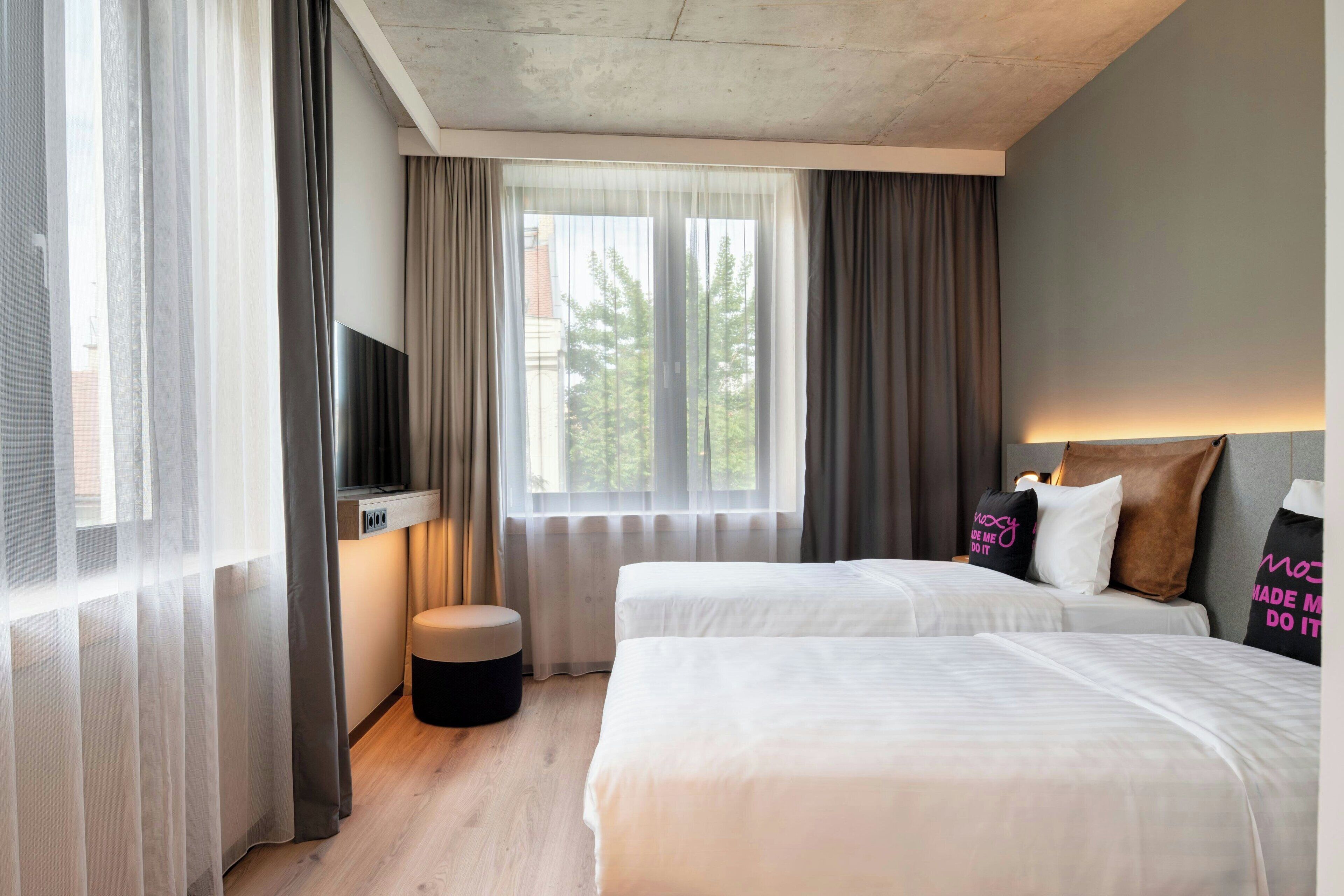 A hotel room with two beds, a TV, a small stool, and a window with curtains.