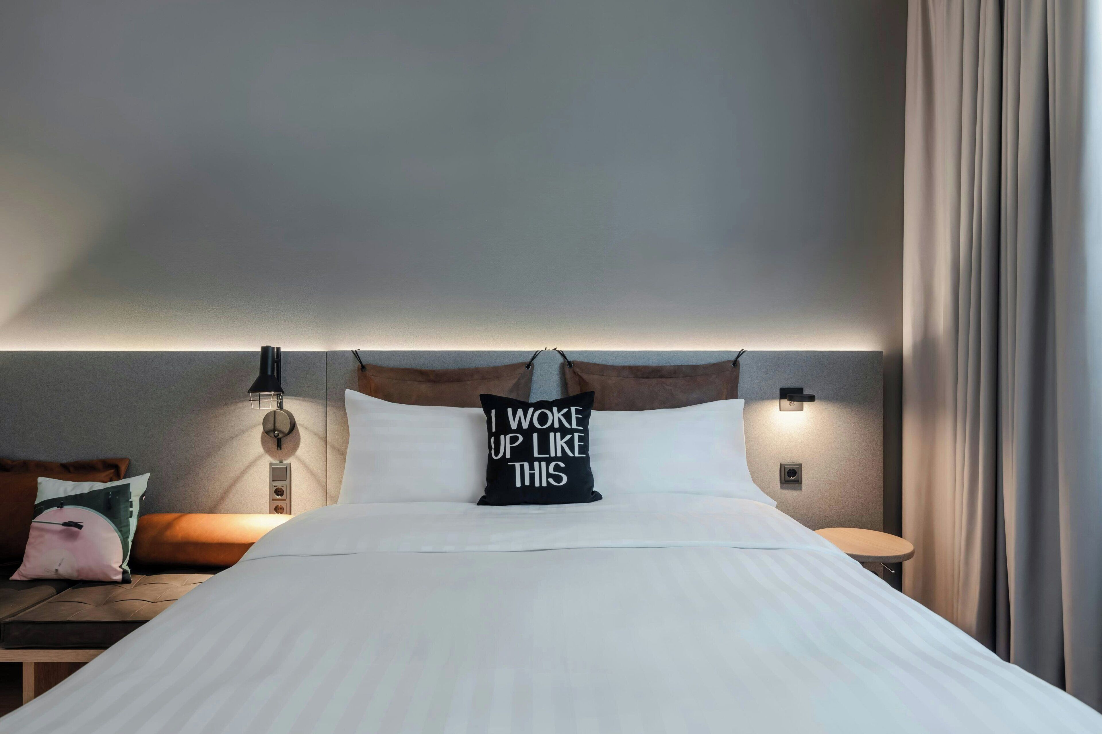 Una cama bien hecha con un cojín que dice 'Me desperté así', una lámpara de noche, una mesita y una cortina.