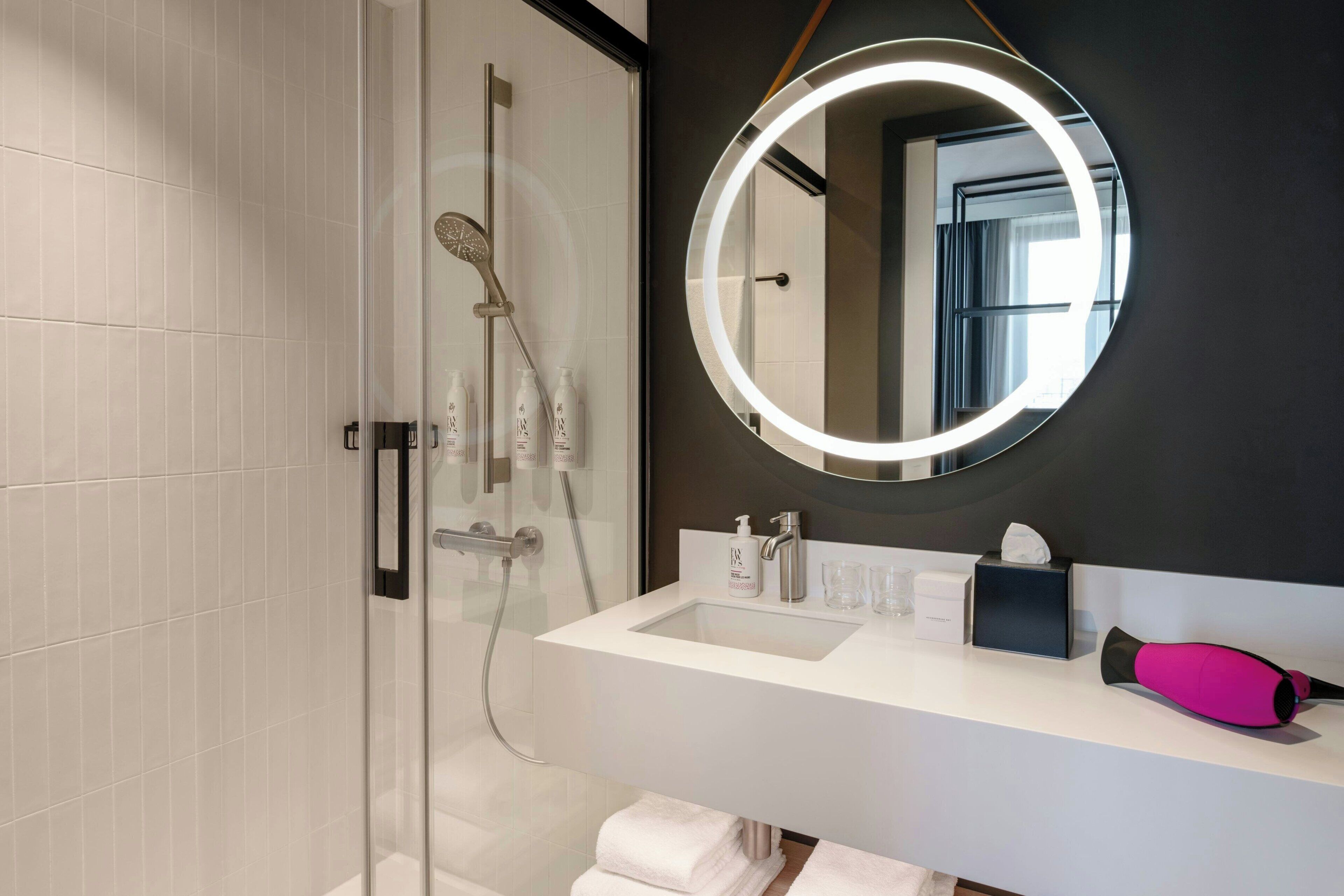 A modern bathroom with a glass shower enclosure, a white sink, a round mirror with a light, and a pink hairdryer.
