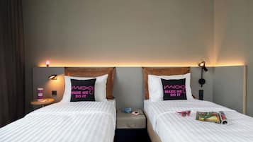 Une chambre d’hôtel avec deux lits, chacun orné d’un oreiller sur lequel est brodé le message « C’est lui/elle qui m’a fait faire ça ». On y trouve des tables de chevet avec des lampes, une petite table avec une tasse et une valise posée sur le sol.