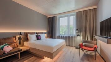 Una habitación de hotel moderna con cama, mesita de noche, silla y un amplio ventanal con cortinas.