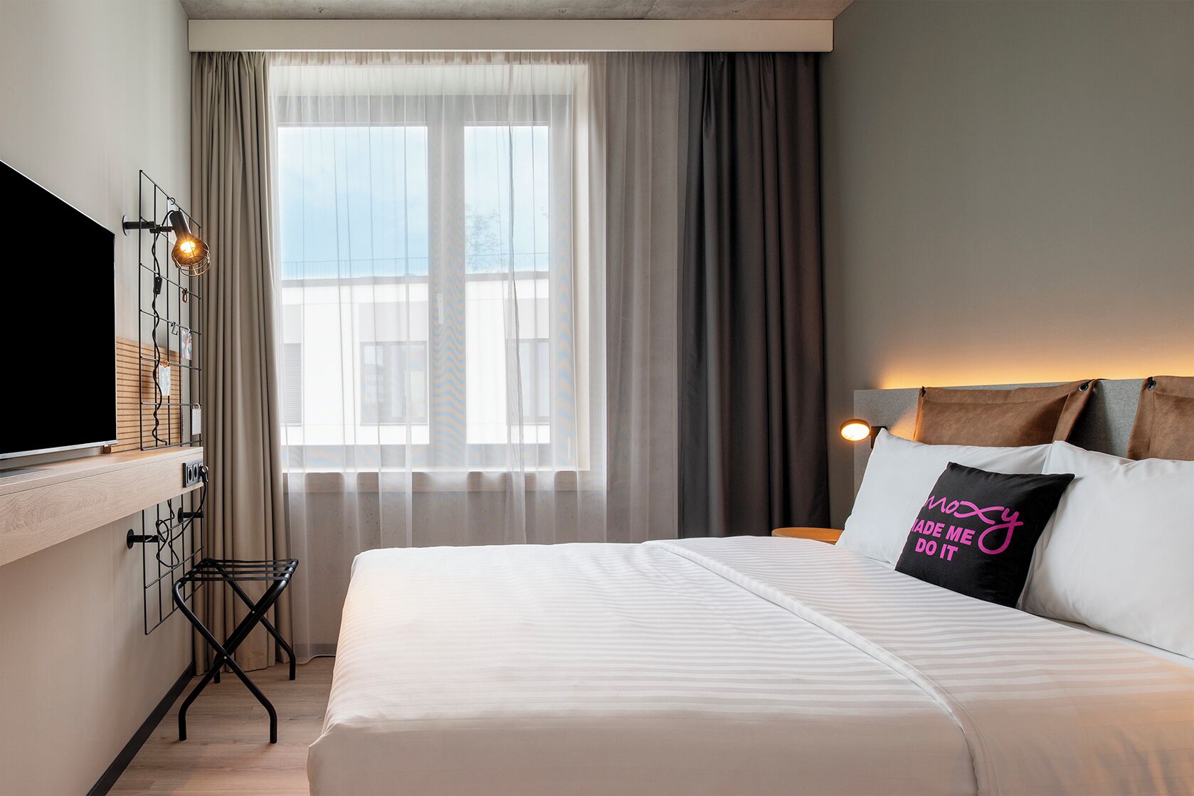 Una habitación de hotel con una cama, un televisor montado en la pared, una ventana con cortinas y una lámpara de noche.