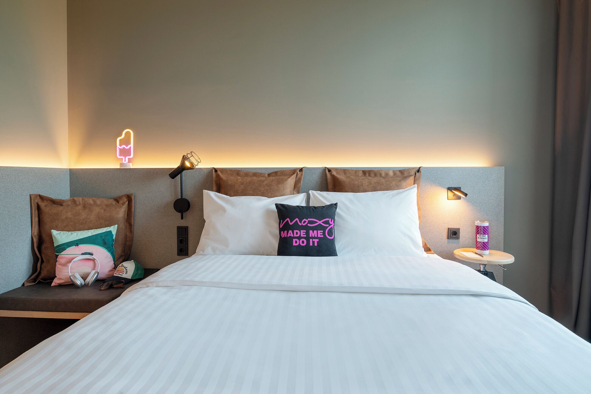 A hotel room with a bed, pillows, a bedside table, a lamp, and a wall-mounted light.
