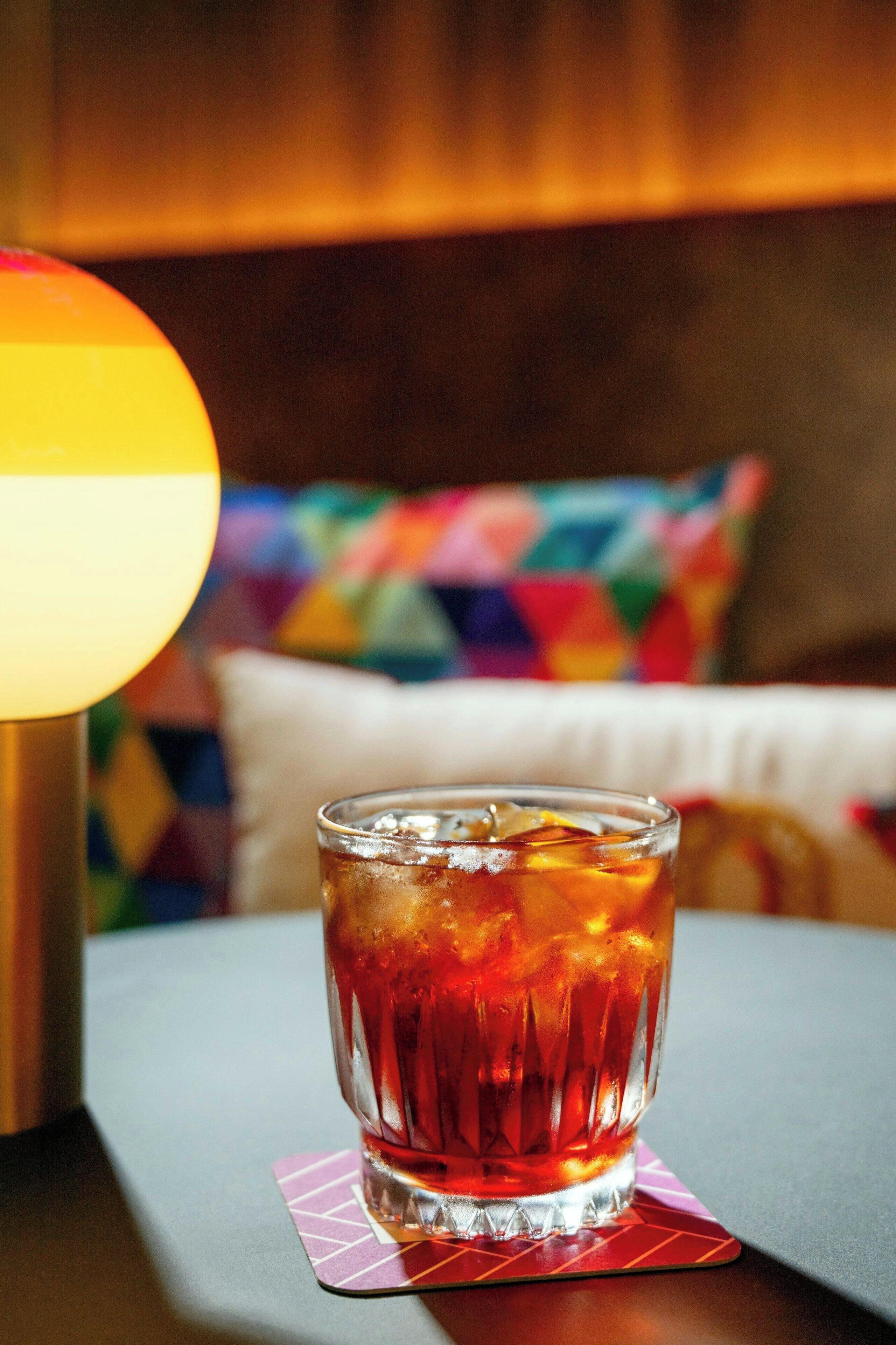 A glass of amber-colored beverage on a coaster, a lamp, and a couch with colorful pillows.