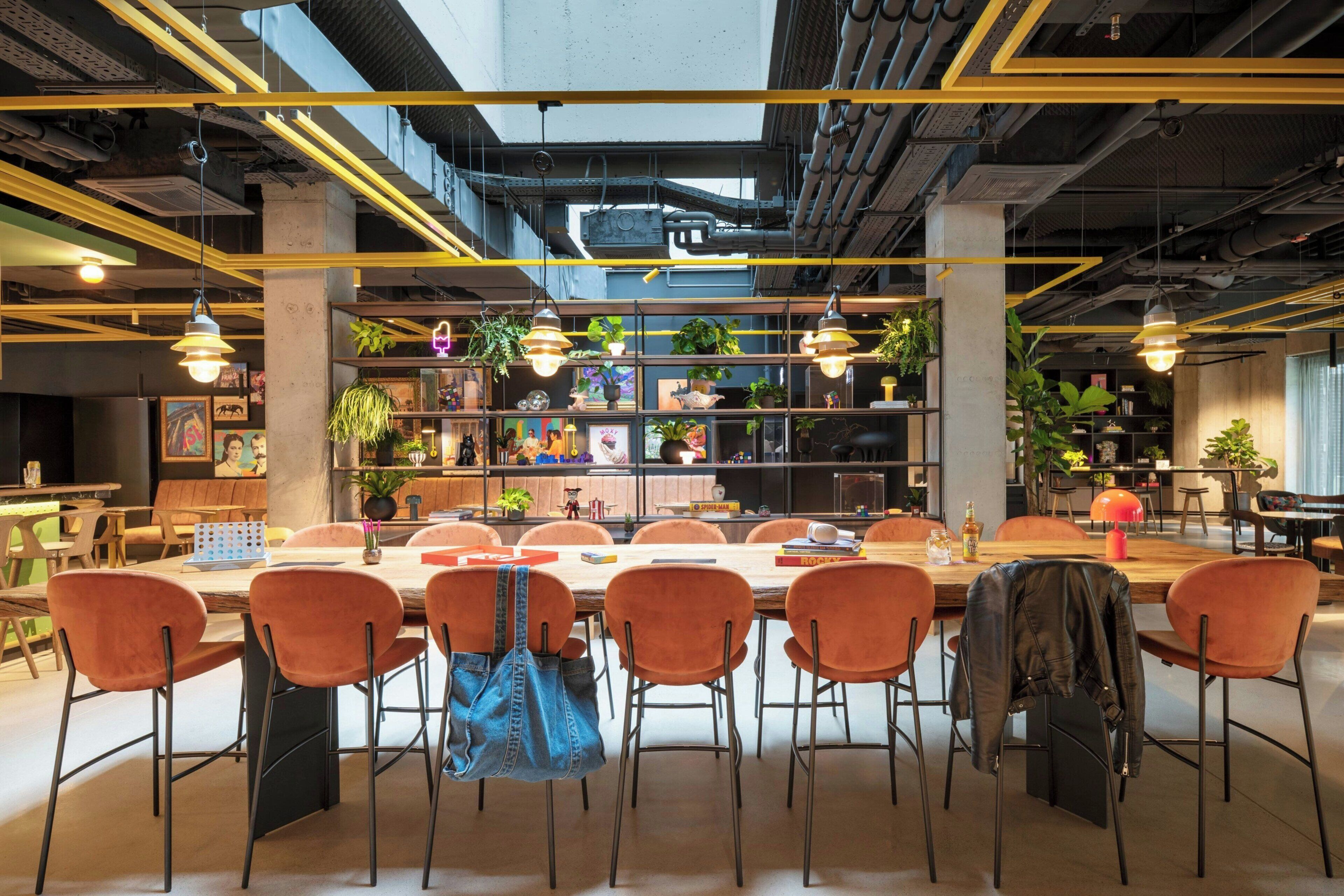 A modern meeting room with a long table, orange chairs, hanging plants, and exposed ceiling pipes.