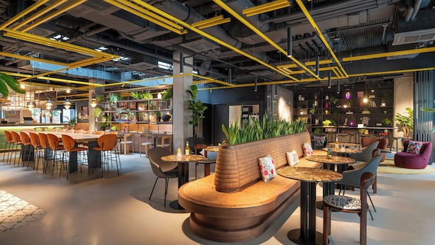 A modern interior with a central seating area, tables, and chairs, surrounded by shelves and plants. - Moxy Budapest Downtown (Budapest)