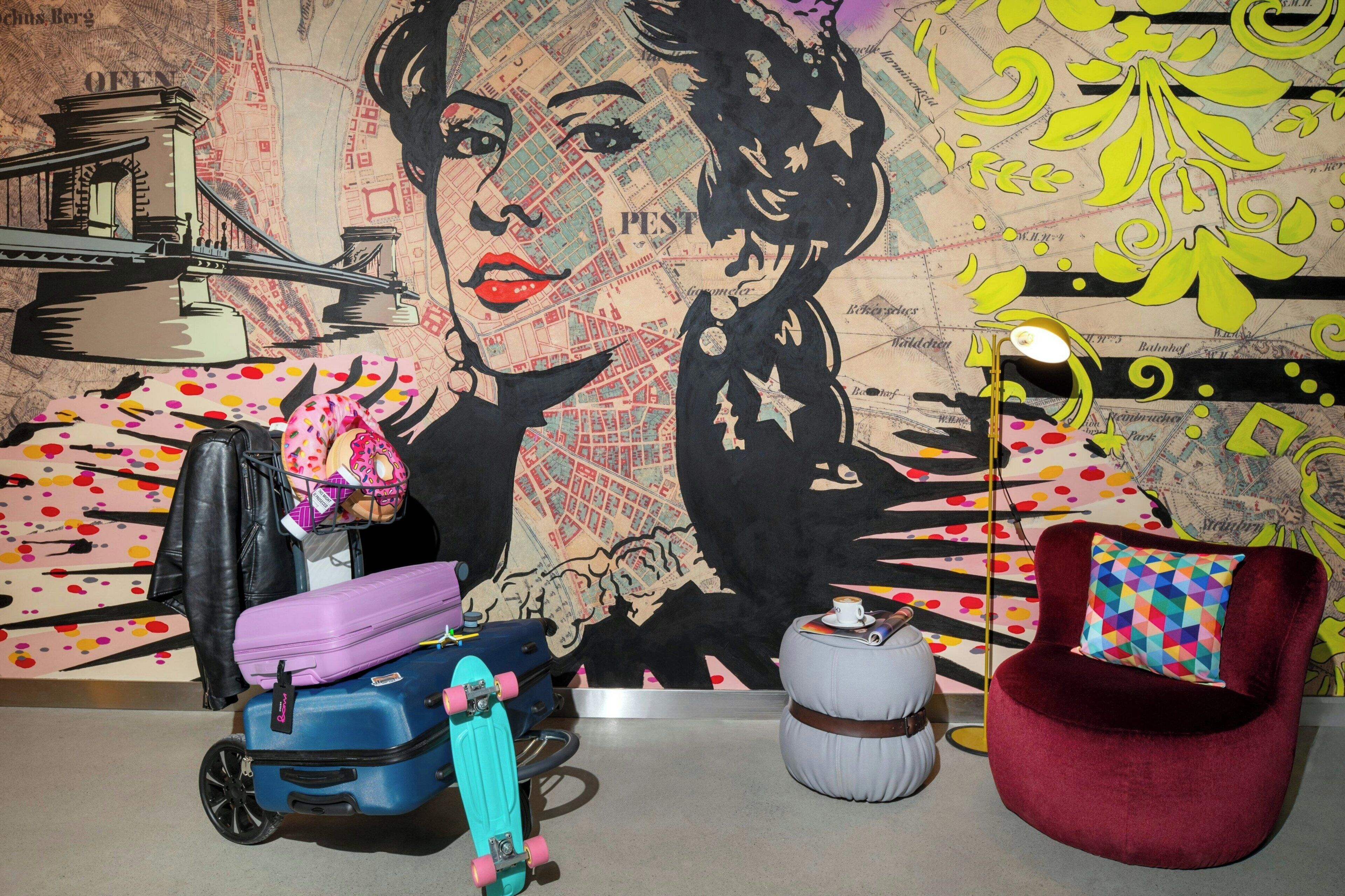A room with a large wall mural, a red chair, a small table, and luggage.