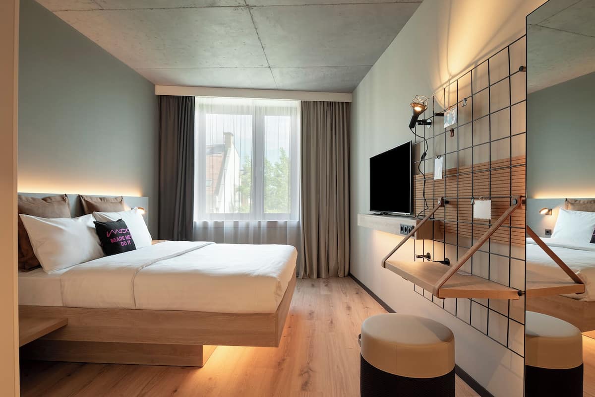 A modern hotel room with a bed, a TV, a wooden bench, and a large window.