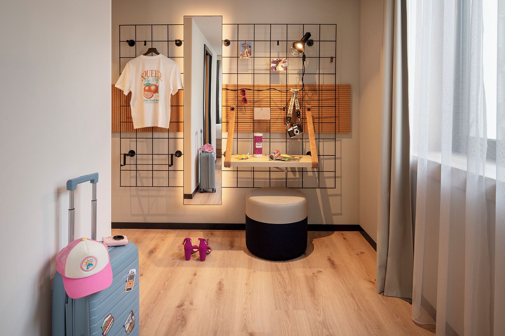 Habitación de hotel con suelo de madera, un escritorio pequeño con espejo, una toalla colgando, una maleta y una gorra rosa.