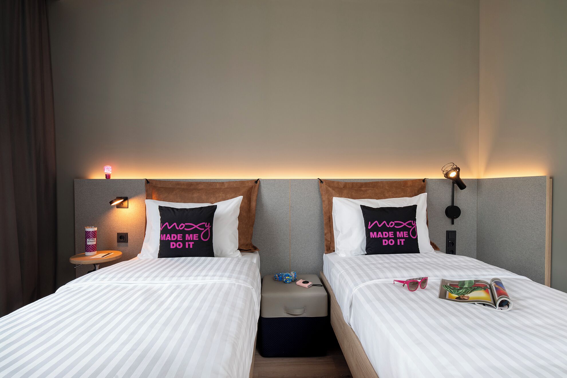 A hotel room with two beds, each with a pillow featuring the text 'Moxy Made Me Do It.' There are bedside lamps, a small table with a candle, and a suitcase on the floor.
