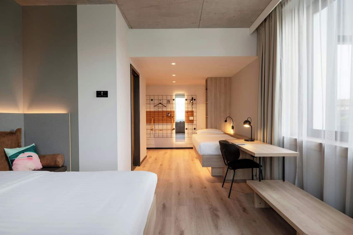 A modern hotel room with a large bed, a desk, and a chair. There is a hallway leading to a bathroom.